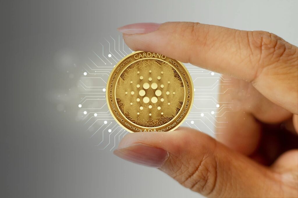 Crypto Cardano in the center, gold color. Held between thumb and forefinger of one hand.